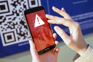 Smartphone showing a red alert warning symbol on screen, with a QR code blurred in the background, representing the risks of QR code scams known as quishing.