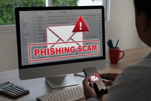 A computer monitor displaying a red alert popup with the text "Phishing Scam," representing a warning about fraudulent emails and DMs attempting to steal personal information.