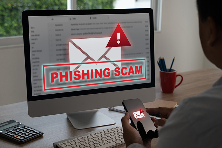 A computer monitor displaying a red alert popup with the text "Phishing Scam," representing a warning about fraudulent emails and DMs attempting to steal personal information.